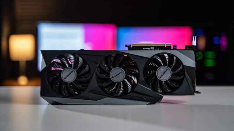 Image shows the Gigabyte RTX 3080 graphics card facing forward on a white desk, with blurred PC screens in the background