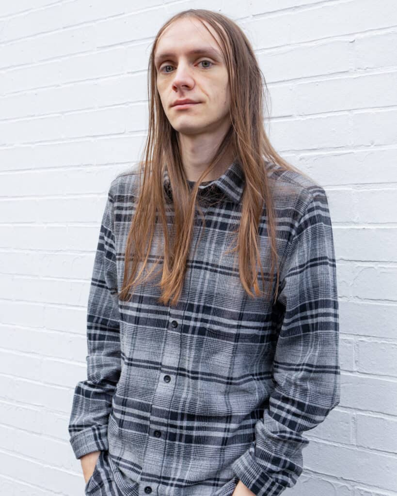 A man with long hair leaning against a white wall.