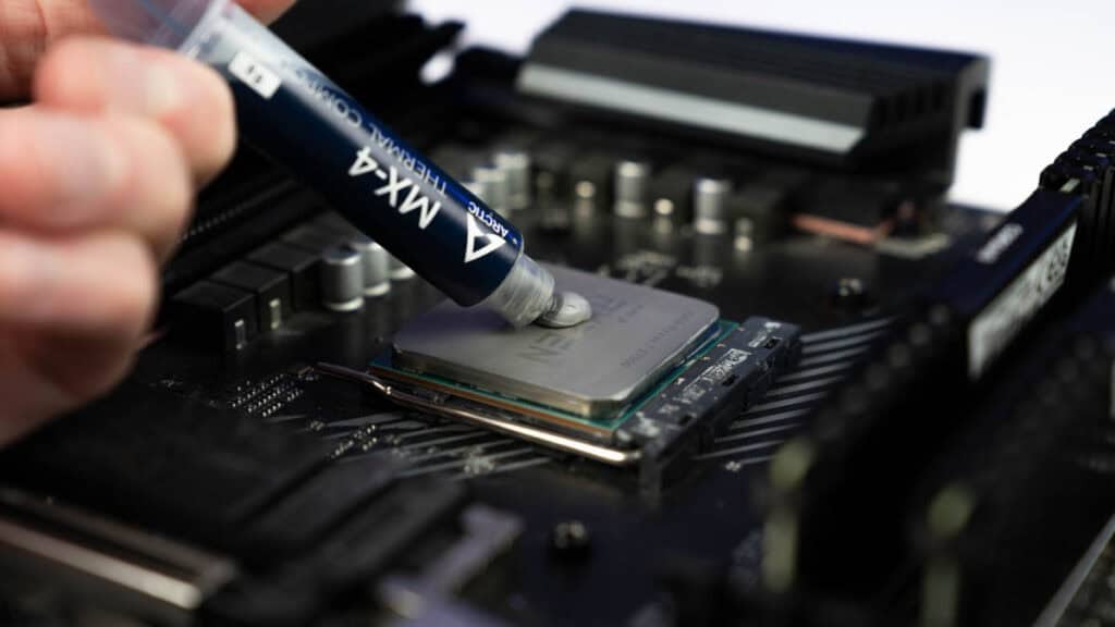 Applying thermal paste from a syringe onto a GPU on a motherboard, surrounded by RAM slots and heat sinks.