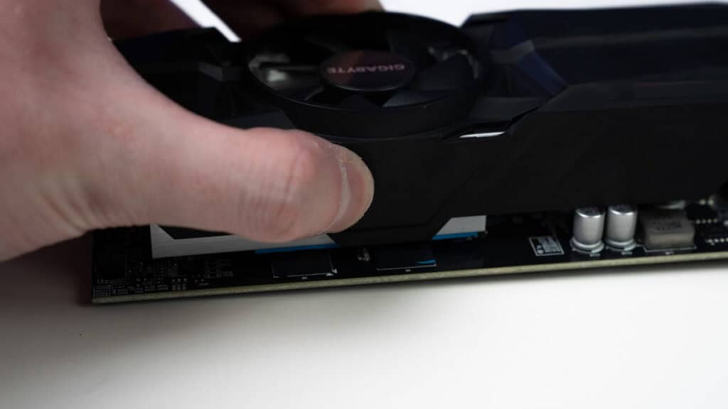 Close-up of a human finger pressing on a black computer graphics card containing one visible fan while demonstrating how to apply thermal paste to a GPU.