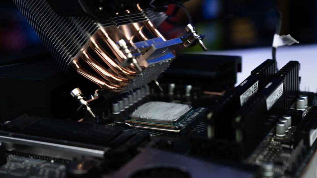 Close-up of a computer motherboard with thermal paste applied on the GPU, surrounded by RAM slots and heat sinks; a robotic arm is positioned above.