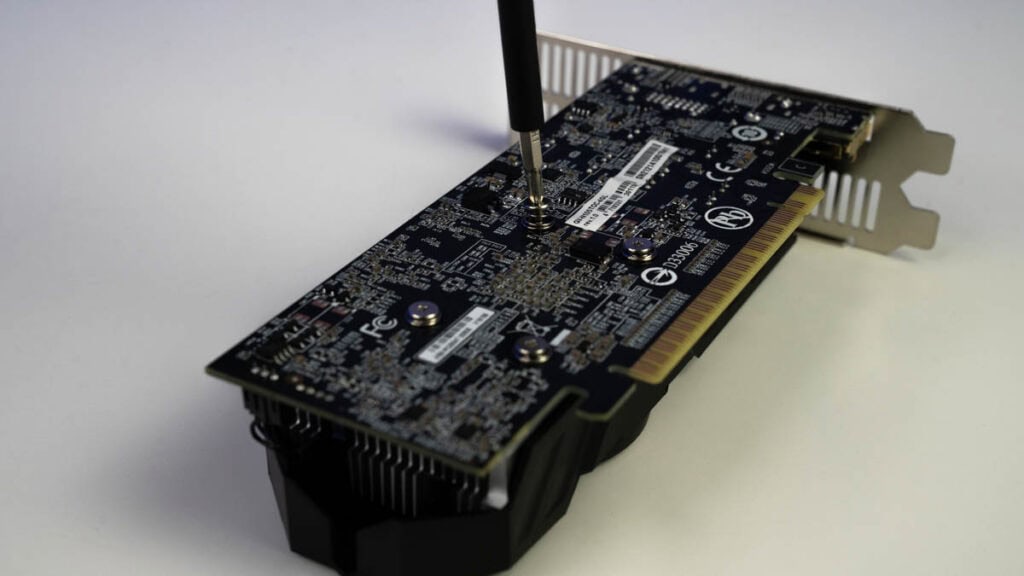 A close-up of a graphics card with a screwdriver adjusting a component on a white background, demonstrating how to apply thermal paste to a GPU.