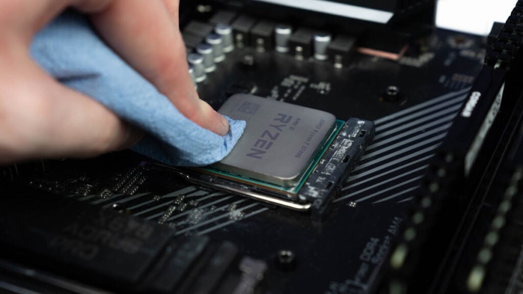 A hand using a cloth to prepare a computer CPU on a motherboard for the application of thermal paste, showing detailed circuit components.