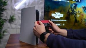 Image shows a person holding a red DualSense controller in front of a PlayStation 5, with a blurred gaming TV in the background. 