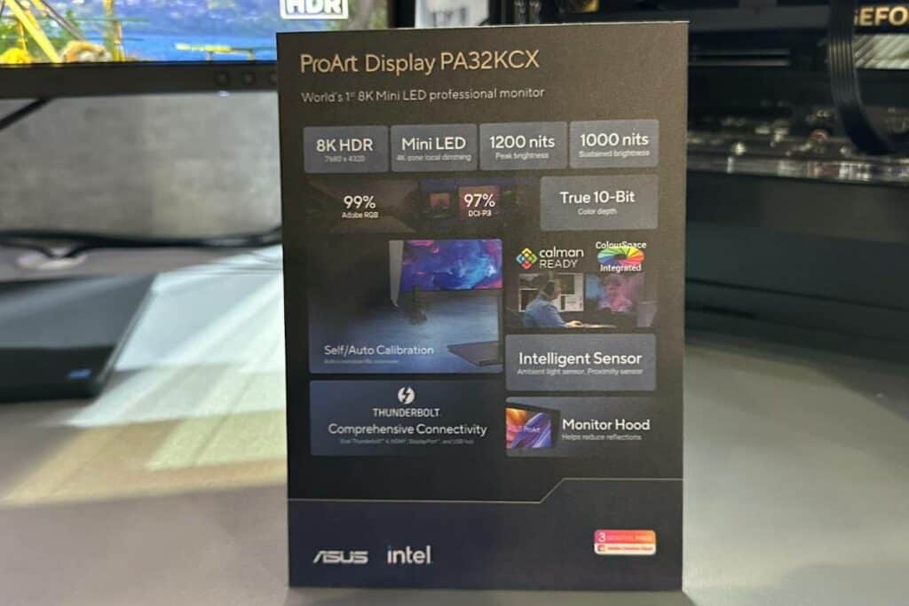 An ASUS PA32KCX display box highlighting features like 8K HDR, mini LED, 1200 nits brightness, and comprehensive connectivity, situated on a blurry desk background.