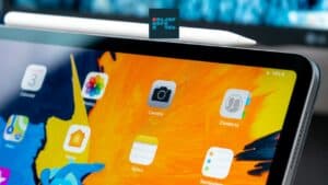Close-up view of an Apple iPad Pro with an orange wallpaper displaying app icons, a digital stylus resting on top, and a 14% battery indicator visible.