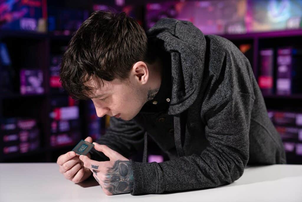 A man with tattoos examines a small electronic device intently in a room with colorful lighting and computer parts, focused on CPU durability, in the background.