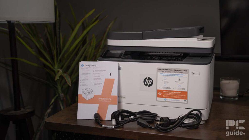 A new HP LaserJet Pro MFP 3102fdwe on a wooden desk with an unfolded setup guide and cables, located next to a small candle, in a room with dim lighting.
