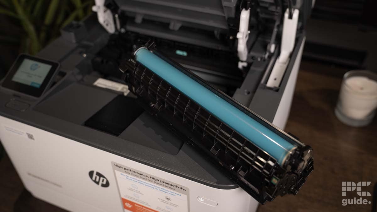 A cyan toner cartridge being installed in an open HP LaserJet Pro MFP 3102fdwe printer, with a small candle and potted plant nearby on a wooden surface.