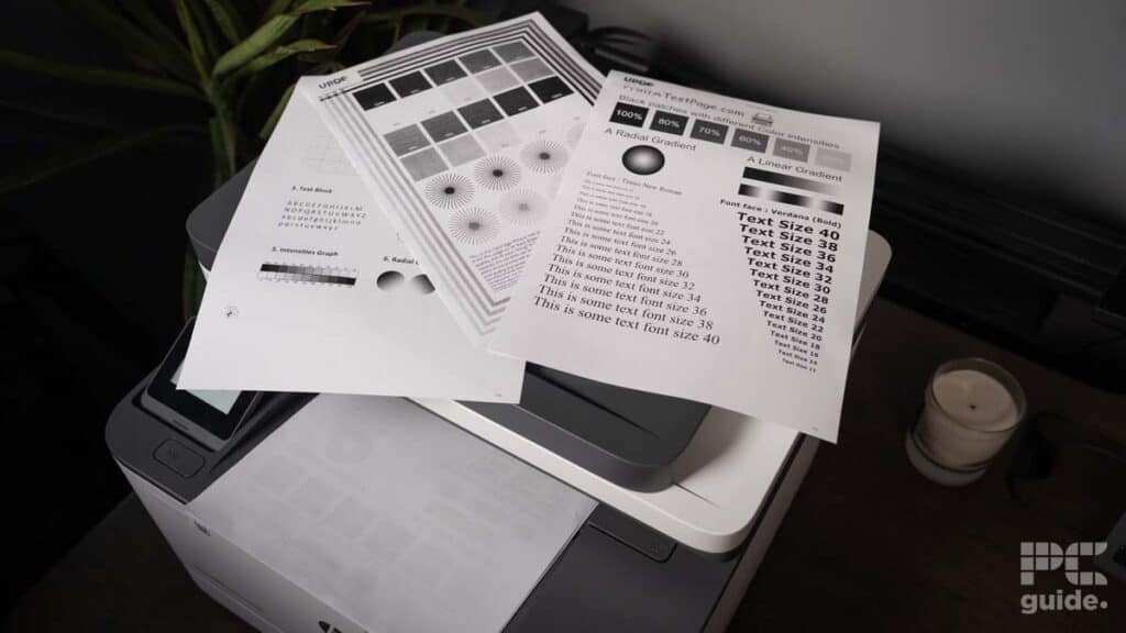 Printed test sheets with various font sizes and patterns lying on top of an HP LaserJet Pro MFP 3102fdwe multifunction printer next to a plant.