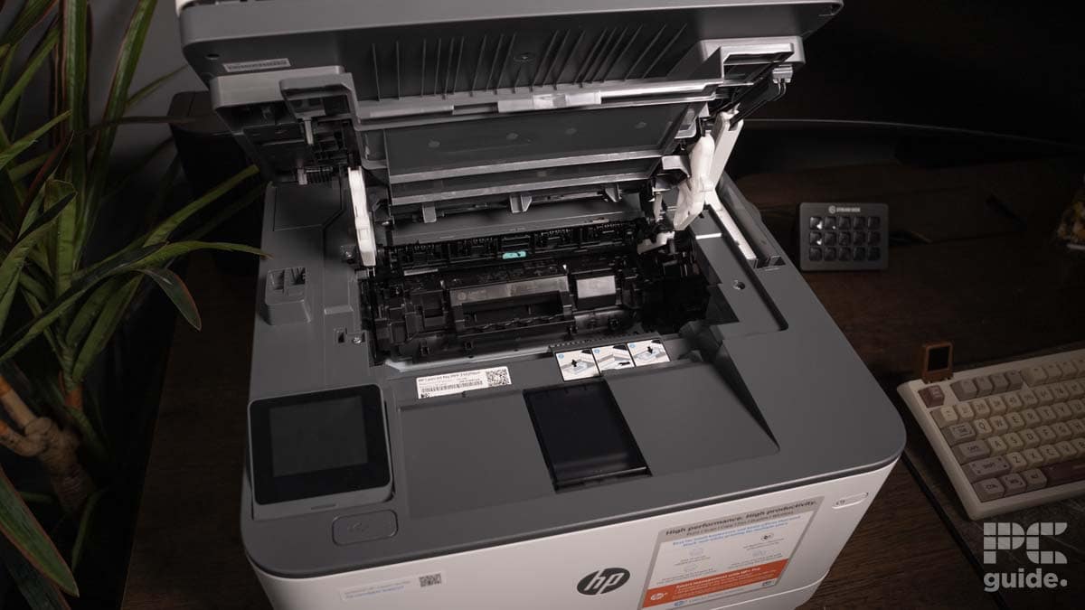 An open HP LaserJet Pro MFP 3102fdwe showing internal components and a paper jam being fixed, with office items and plants in the background.