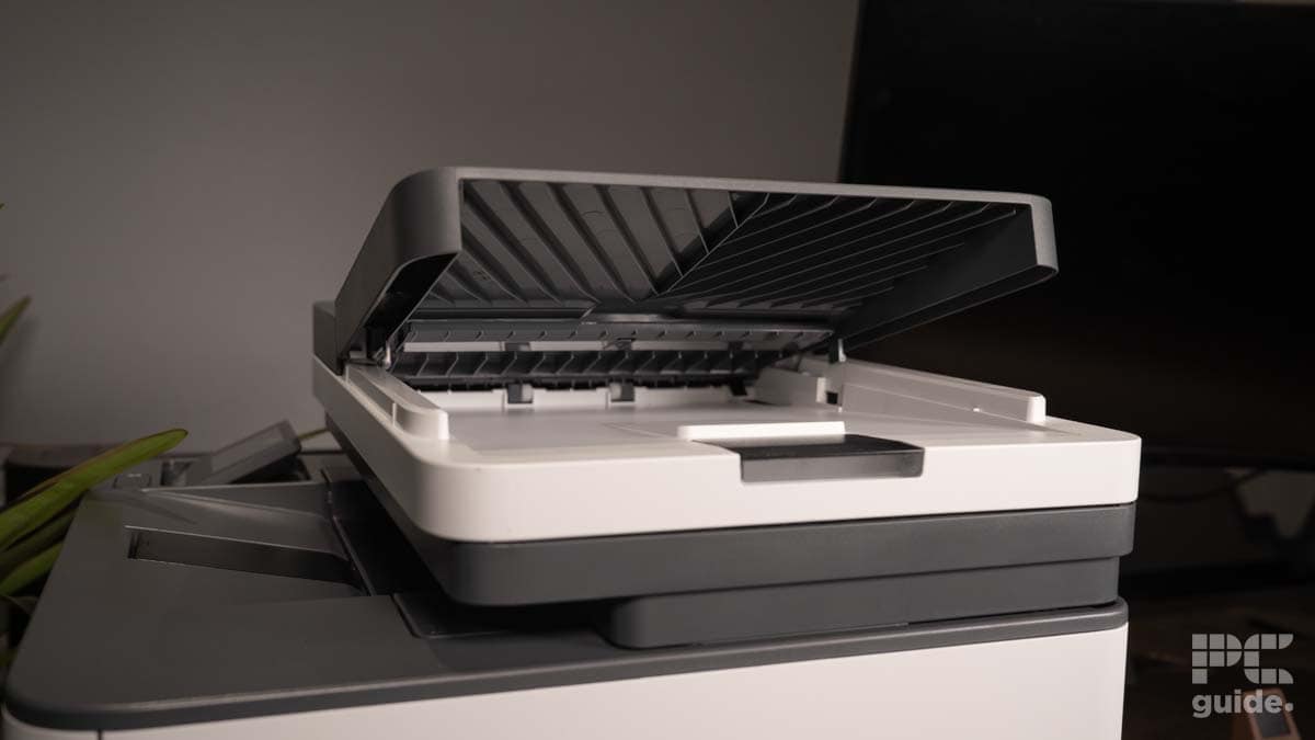 A modern HP LaserJet Pro MFP 3102fdwe sits on a desk with its top cover open, showing the internal paper tray, set against a soft-lit backdrop.