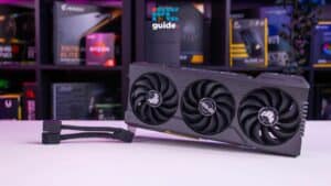 A triple-fan graphics card sits on a table with colorful, lit PC components in the background, showcasing how to enable Hardware Accelerated GPU Scheduling. Image taken by PCGuide.com