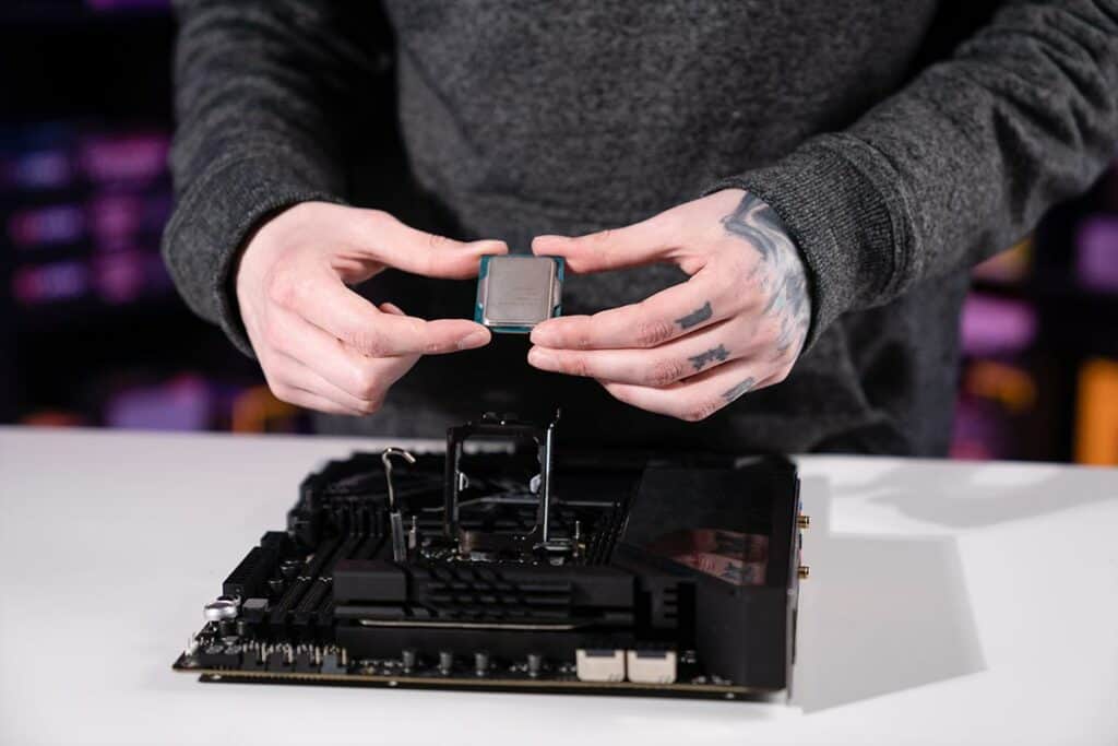 A person holds a CPU chip above a motherboard, preparing to install it amid signs of CPU problems. The background is softly blurred with purple lighting.