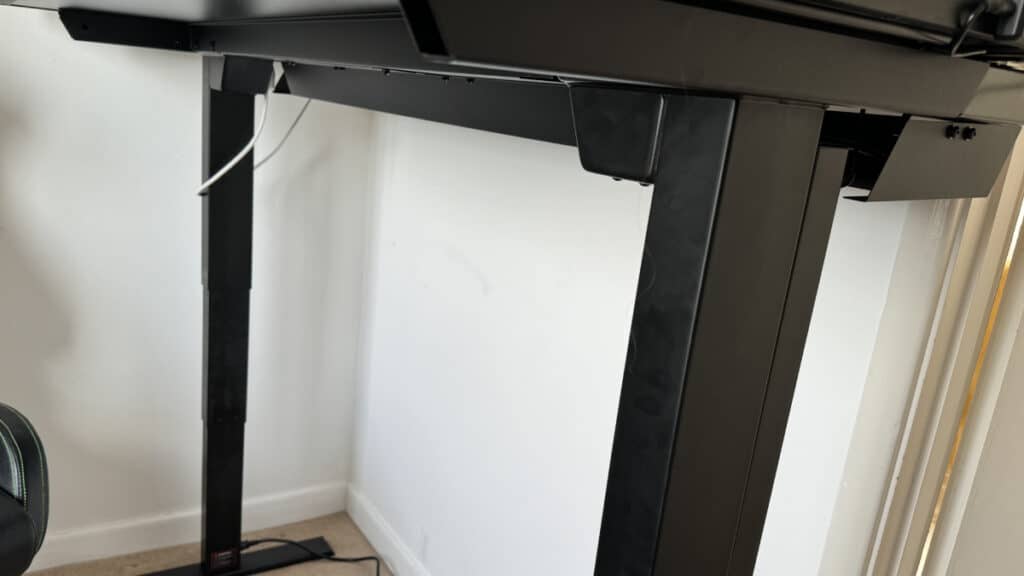 A black adjustable Secretlab Magnus Pro standing desk with visible metal legs in a white-walled room, showing a portion of a chair on the left.