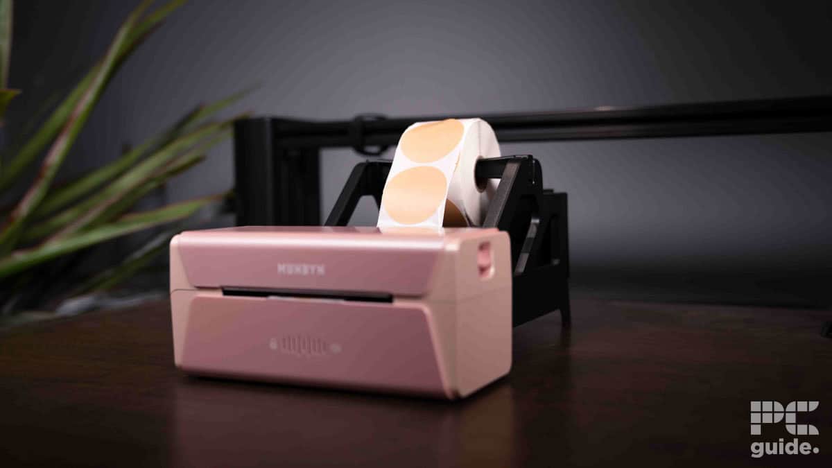 A pink Munbyn RW401AP label printer on a desk, processing a roll of vinyl stickers, with a plant in the background.