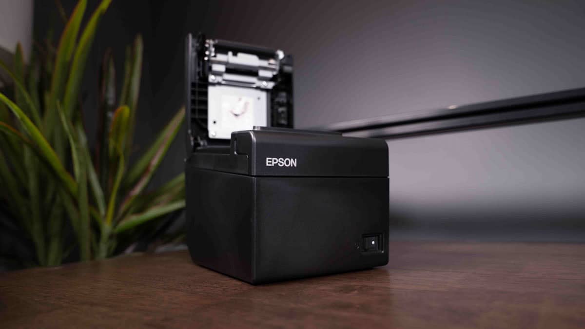 An Epson TM-T20II thermal receipt printer with an open lid on a wooden desk, next to a potted plant and under soft lighting.