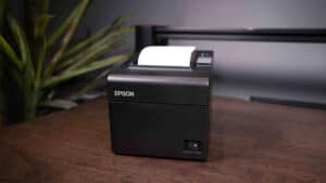 An Epson TM-T20II thermal receipt printer on a wooden desk next to a potted plant.