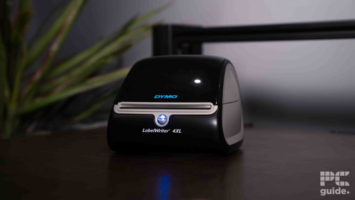 A black Dymo LabelWriter 4XL label printer on a wooden desk, with a potted plant and a monitor in the softly lit background.