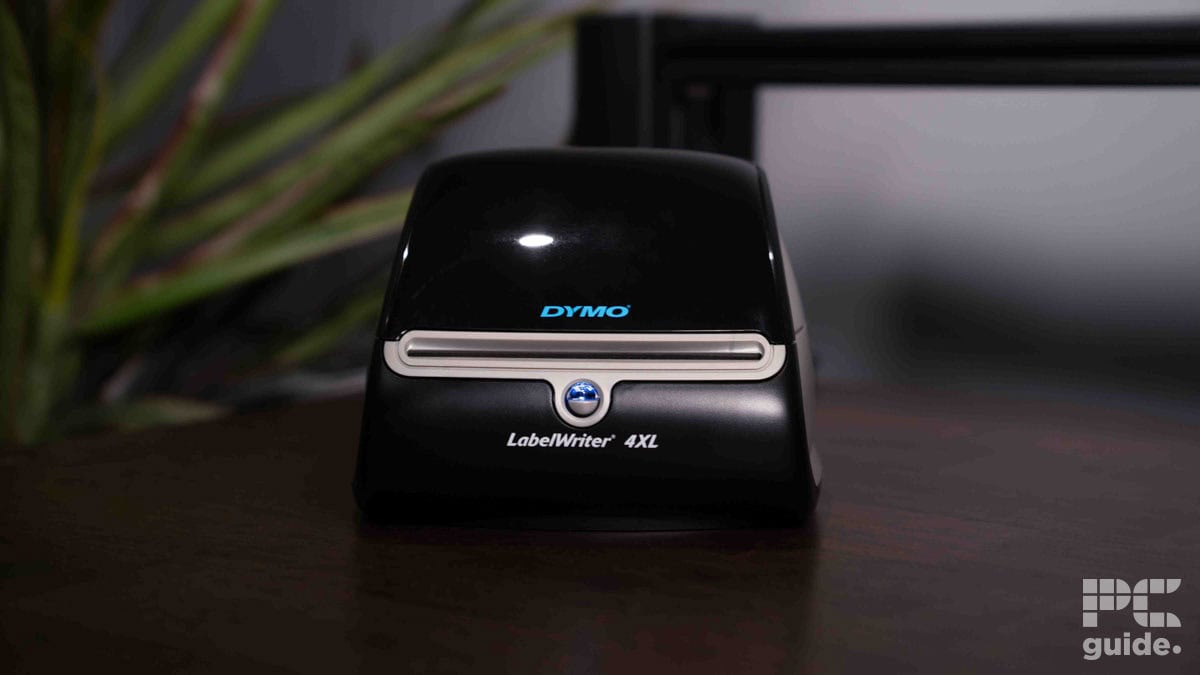 A black Dymo LabelWriter 4XL thermal label printer on a desk next to a potted plant, with soft focus on the background.