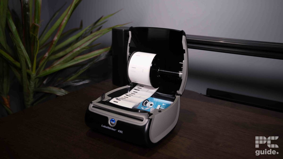 A Dymo LabelWriter 4XL label printer open with a roll of labels on a wooden desk beside a potted plant, with a "ps guide" logo visible on the image.