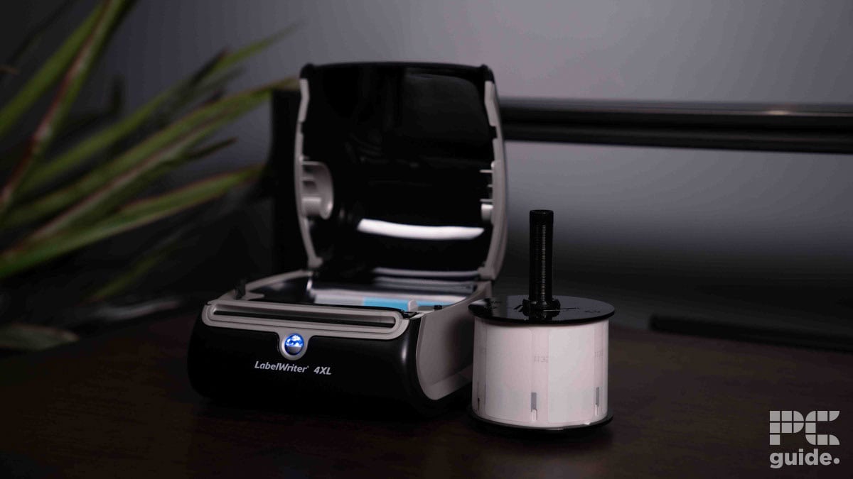 A Dymo LabelWriter 4XL label printer on a desk next to a plant, with a roll of labels partially inserted into the printer.