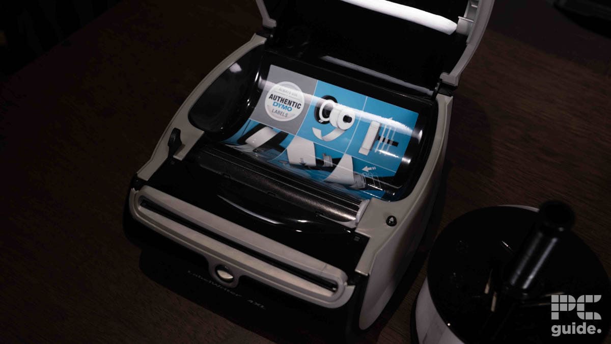 A Dymo LabelWriter 4XL desktop label printer with an open lid and a sheet of paper featuring the Google logo halfway through printing, positioned on a dark wood surface.