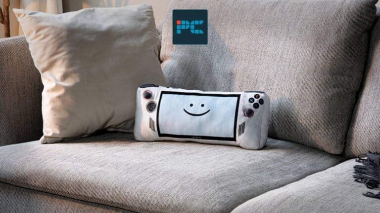 A plush toy resembling a handheld game console with a smiling face is placed on a gray upholstered couch with a cushion, delighting handheld players.