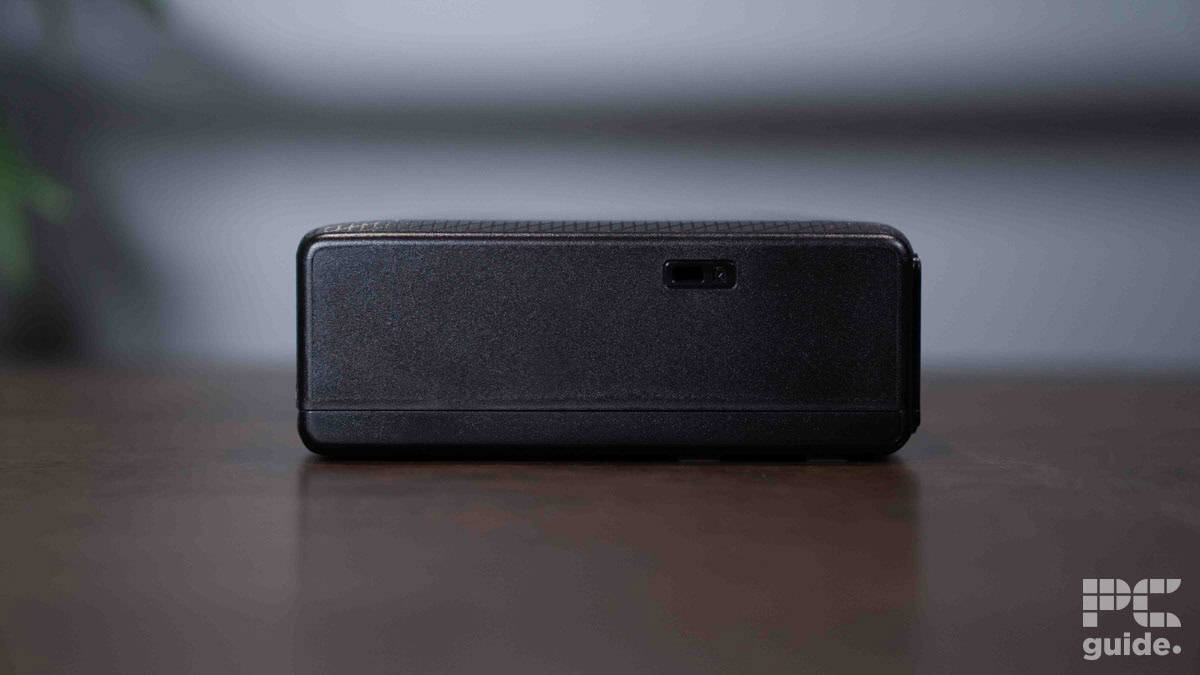 A close-up of a black, rectangular electronic device on a table. The device, resembling an Epson WorkForce WF-110 compact printer, has a small port on one side with the "PC guide." logo in the bottom right corner.