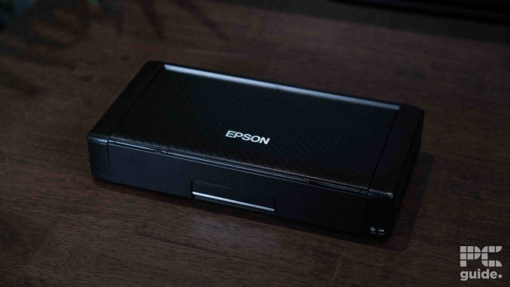 A closed black Epson WorkForce WF-110 portable printer sits on a wooden surface, with the brand name centered on the top. Compact and light, the device features the PC guide logo in the bottom right corner.