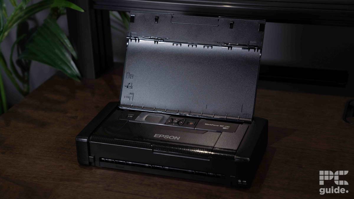 A photo of an Epson scanner with its lid open, placed on a wooden table. A plant is visible in the background, along with part of an Epson WorkForce WF-110 Printer and a monitor.