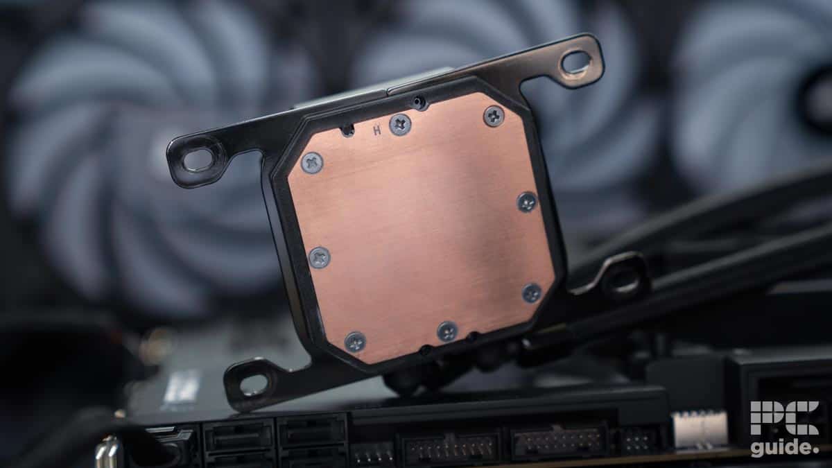 Close-up view of the baseplate of a Corsair CPU cooler, showing a square copper surface with screws along the edges. The background includes blurred computer components and the "PC guide" logo in the corner, highlighting the ELITE LCD on an H150i model.