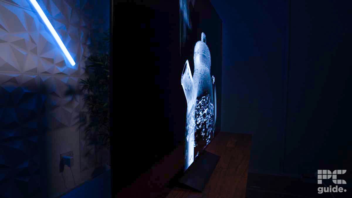 A side view of a sleek, thin television displaying a close-up image of a bubble, reminiscent of an LG C4 OLED review. The room is dimly lit with a blue LED light on the left wall. PC guide™ logo is in the bottom right corner.