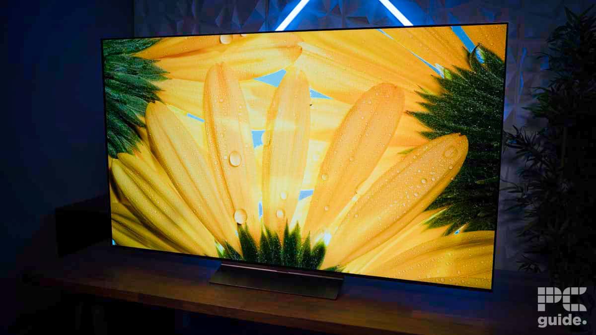 A widescreen TV displays a close-up image of yellow flower petals with water droplets. The TV, subject of an impressive LG C4 OLED review, is placed on a wooden surface with a neon light in the background.