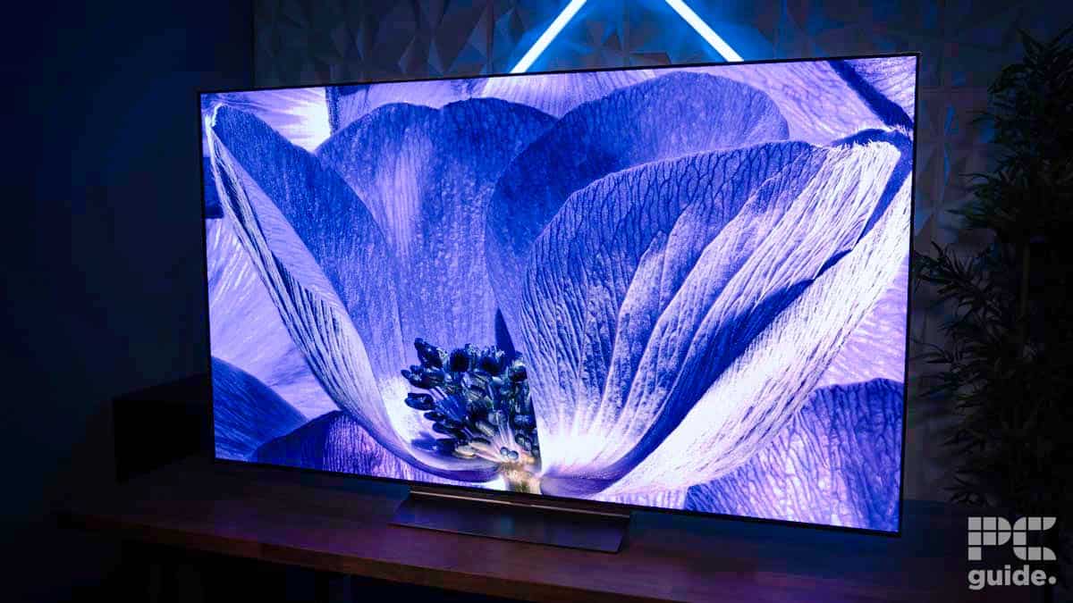 A flat-screen television with a vivid blue and purple flower displayed on the screen, placed on a wooden surface with a lit geometric pattern on the wall behind. The PC Guide logo is in the corner, showcasing our latest LG C4 OLED review.