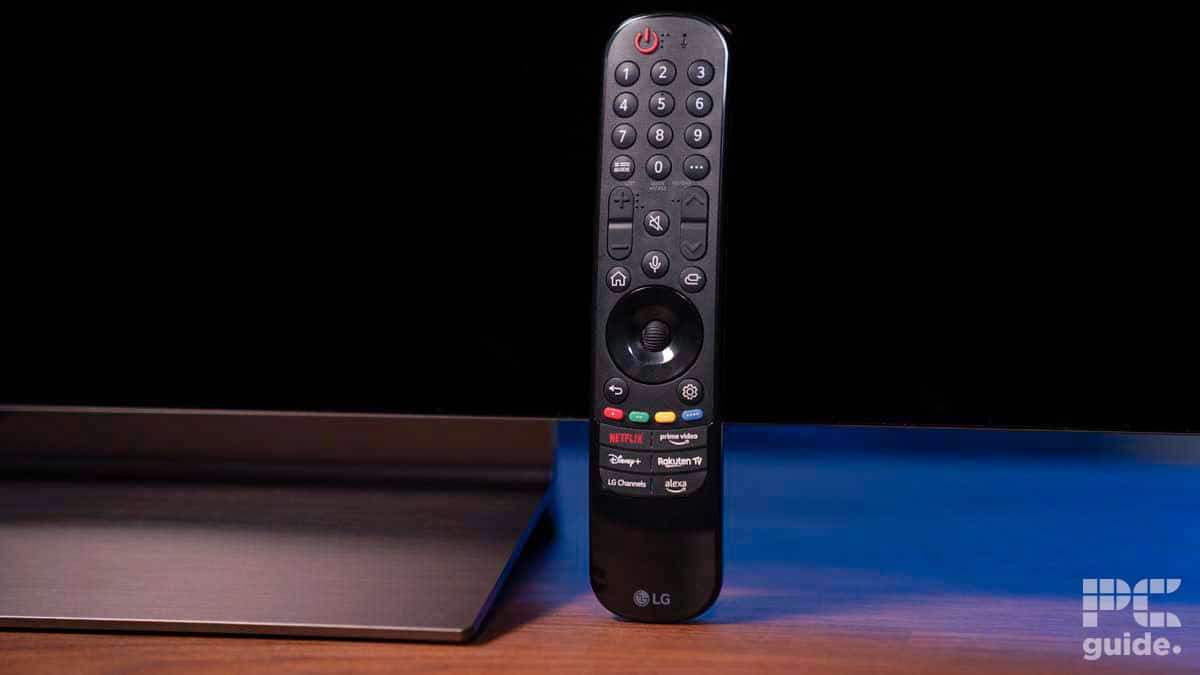 A black LG remote control is standing upright on a wooden surface, with the PC Guide logo visible in the bottom right corner. The background is dark with a hint of a screen on the left, reminiscent of an LG C4 OLED review setup.