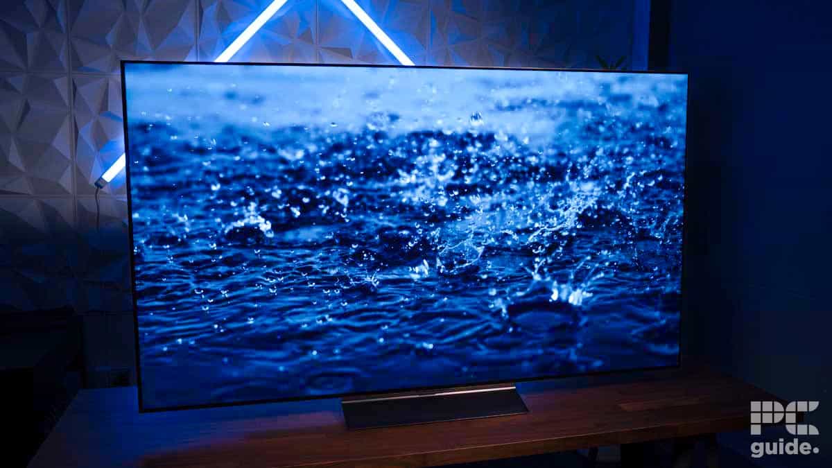 A flat-screen LG C4 OLED TV displaying a close-up image of water droplets splashing on a surface. The TV is placed on a wooden table with blue LED lights in the background, perfect for those considering an LG C4 OLED review.