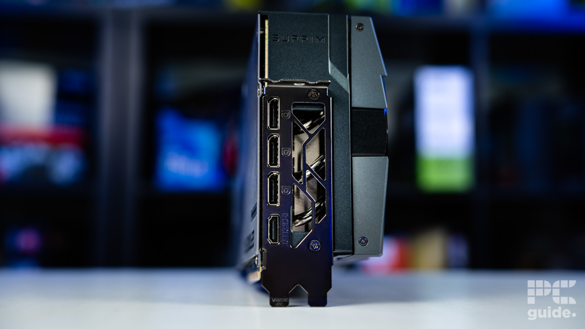 A background of blurred tech boxes sitting on bookshelves. The MSI Suprim SOC RTX 5090 sits on top of a white table. The card is end on showing its video outputs. There are thre DisplayPort and one HDMI. Above it there is lettering reading SUPRIM.