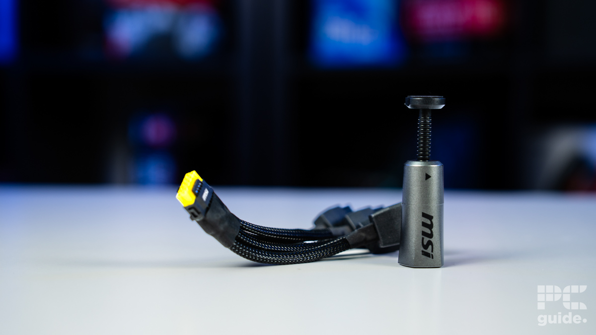 A background of blurred tech boxes sitting on bookshelves. On the white table in the foreground is a 600W 12-pin adapter with yellow connectors for the 5090 and a support stand.