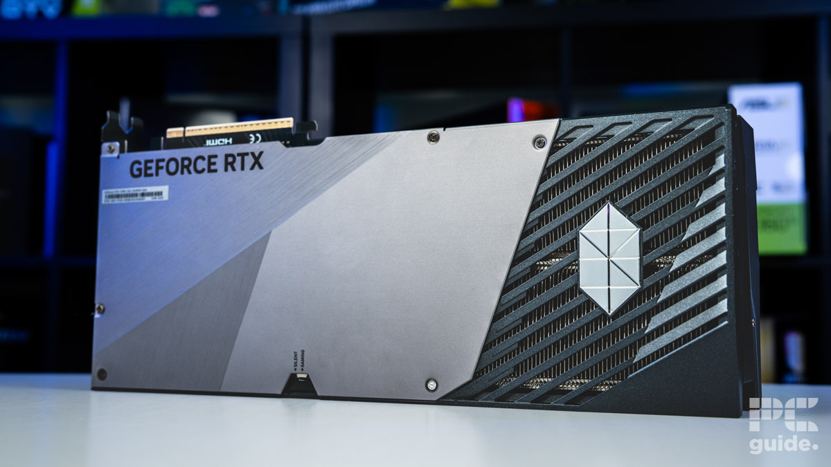 A background of blurred tech boxes sitting on bookshelves. The MSI Suprim SOC RTX 5090 sits on top of a white table. It's a shot of the backplate that is majority silver metal with the right end bineg plastic flow cut out for flow through with the diamond emblem.