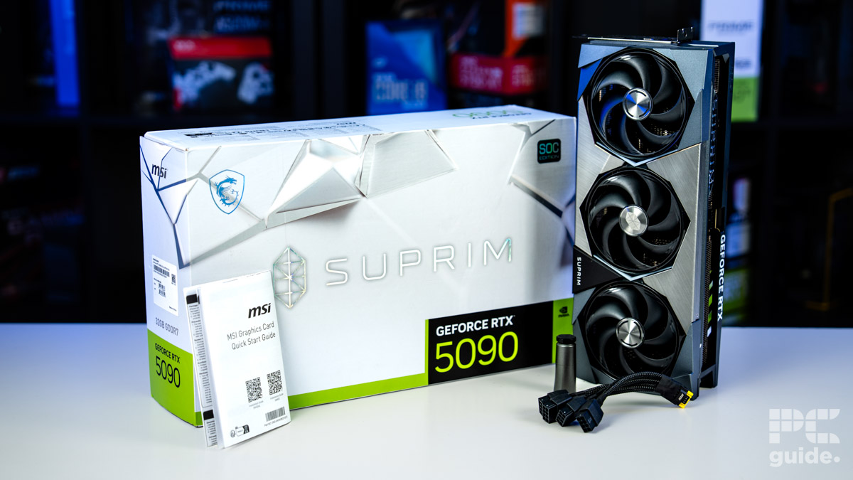 A background of blurred tech boxes sitting on bookshelves. The MSI Suprim SOC RTX 5090 stands verticallt on top of a white table. It's next to the white box with the MSI, Suprim, and RTX 5090 logos. Along with the start guide booklet, connector adapter, and support stand.