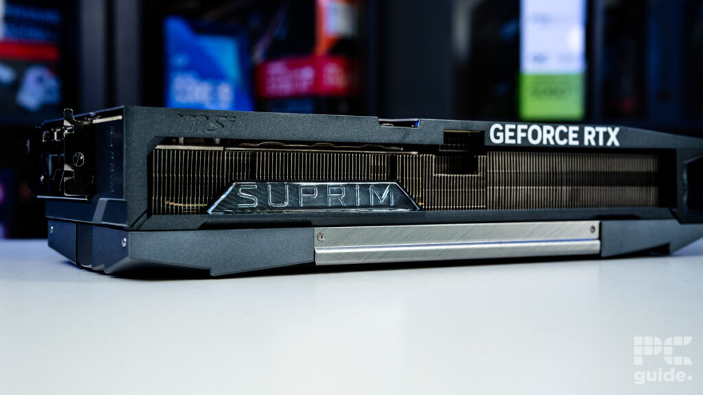 A background of blurred tech boxes sitting on bookshelves. The MSI Suprim SOC RTX 5090 sits on top of a white table. It's a shot of the side edge that shows the big heatsink inside, along with the logos for MSI, SUPRIM, and GeForce RTX.