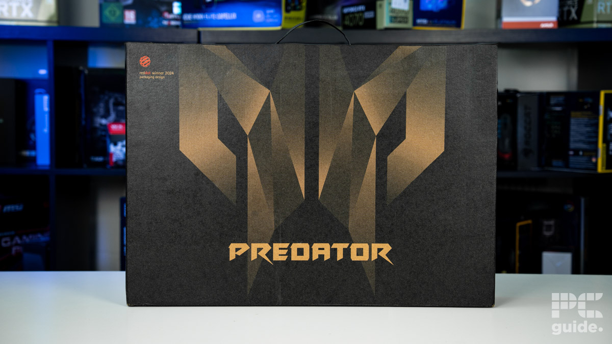 The box for the laptop with the predator logo covering it, and it reads Predator towards the bottom. Sitting on a white table with a blurred background of shelves filled with boxed of tech products.