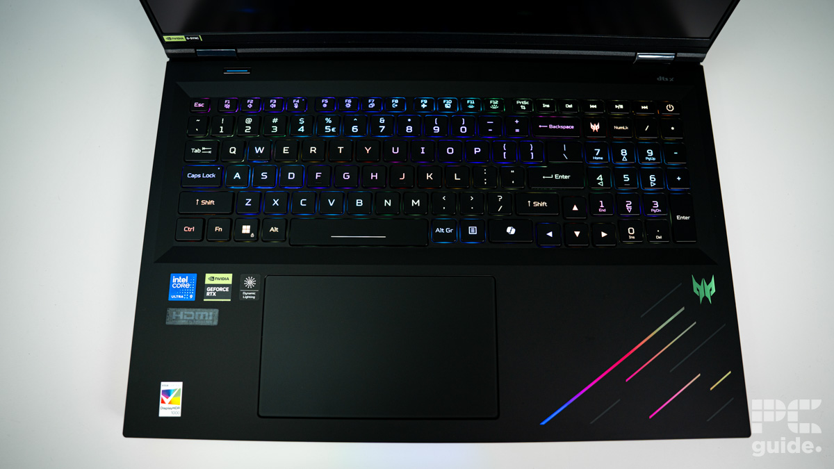 A black laptop shot of the keyboard and touchpad. It is lit up in RGB for the keys, with a range of specs stickers filling the bottom left and RGB strips on the right.