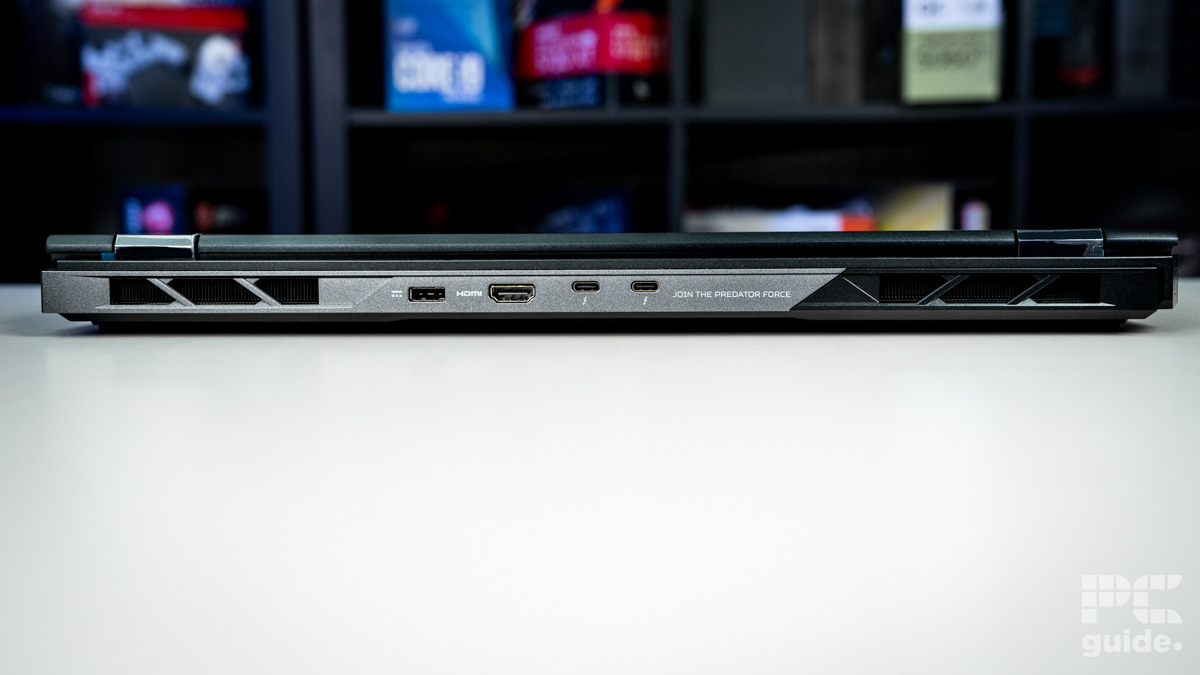 The back ports of the black laptop. On the left and right are vents with metal support bars cutting through. IN the middle are the ports, which from left to right are: charger port, HDMI, and two USB-C. Sitting on a white table with a blurred background of shelves filled with boxed of tech products.