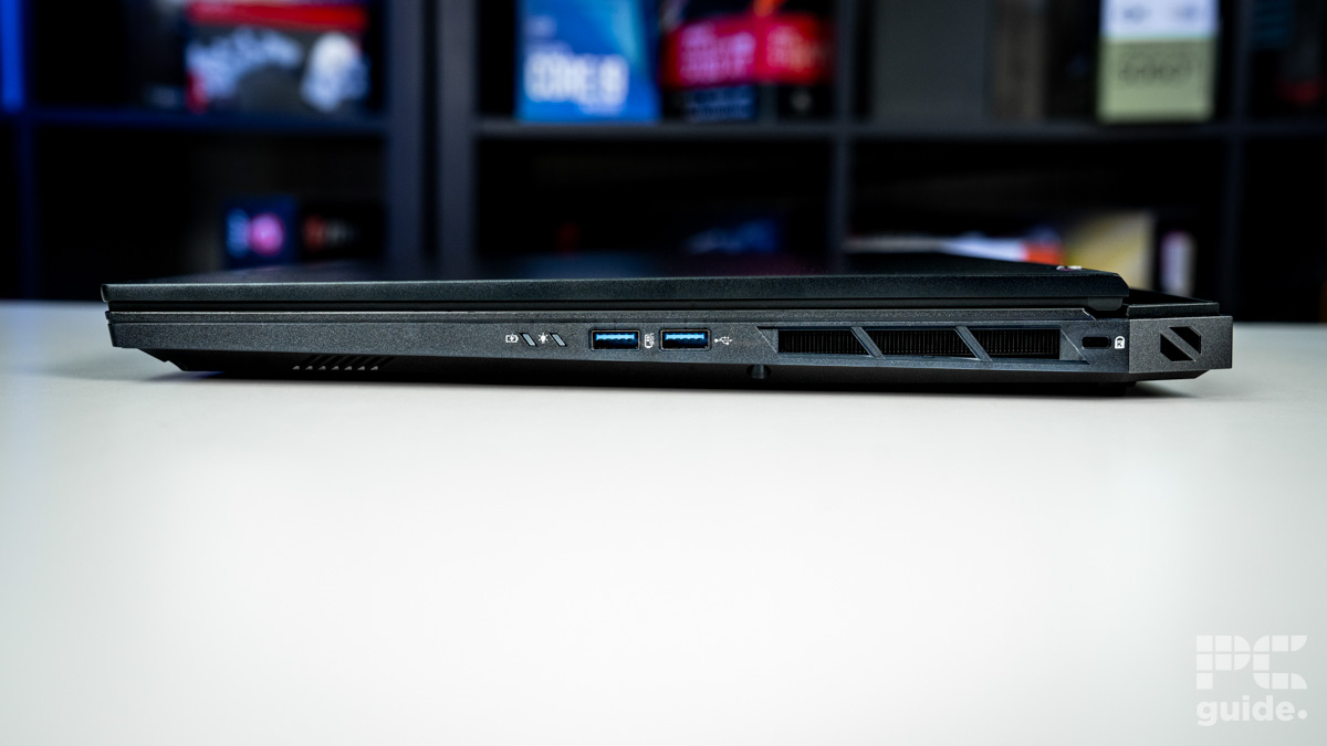 Right side of the balck laptop. On the right is a vent with a kensington lock. In the middle are two USB A ports and two indicator lights. Sitting on a white table with a blurred background of shelves filled with boxed of tech products.