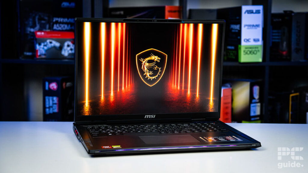 A blurred background of a bookshelf filled with blurred boxes of tech products, with a white table in the foreground. On it sits the black MSI Raider laptop open adn turned on. As the background it has a gold MSI logo on black background with red lights cutting up and down either side of it.