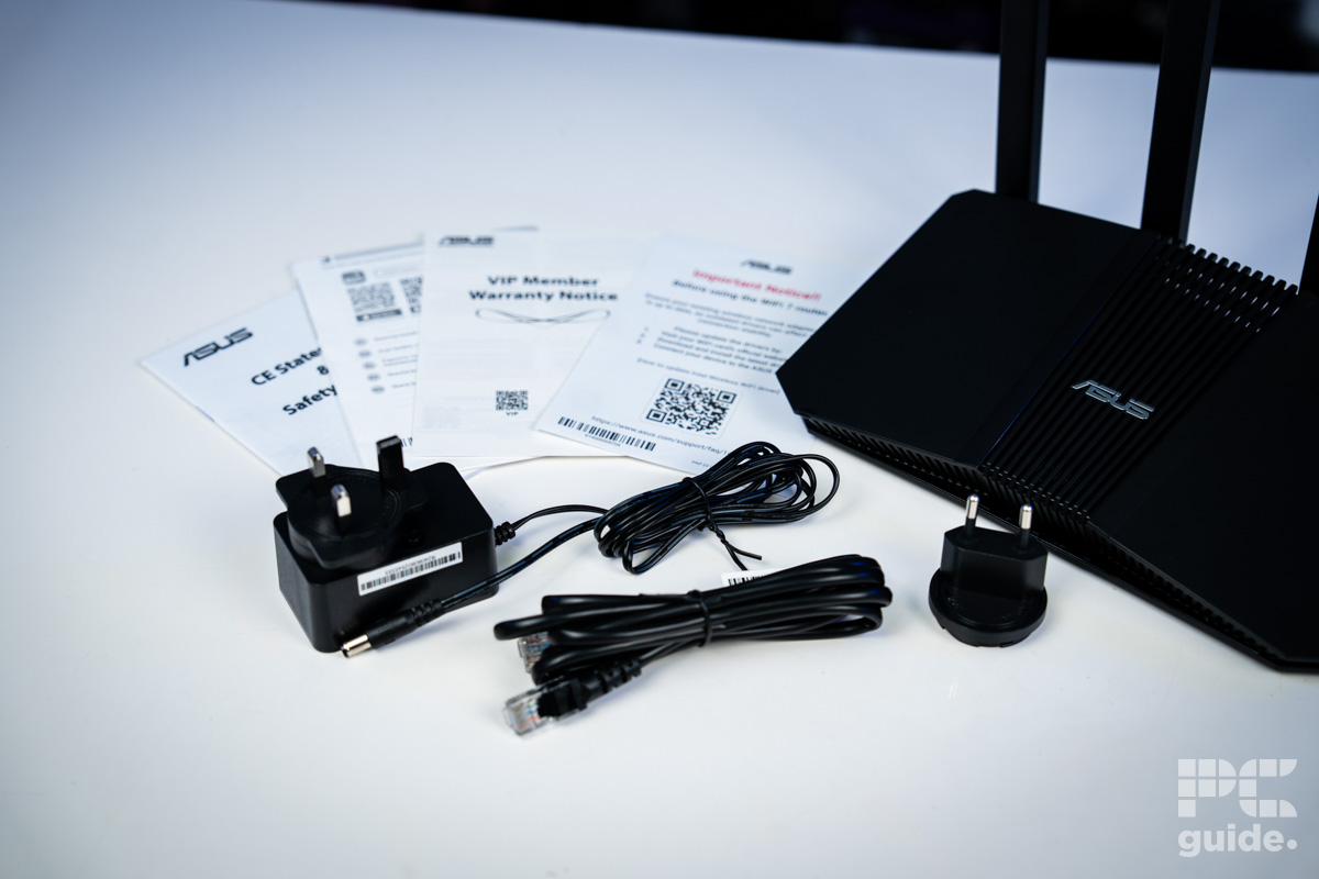 ASUS RT-BE58U sitting on a white table. Next to it is the power adapter, ethernet cable, alternative adapter head, and leaflets.