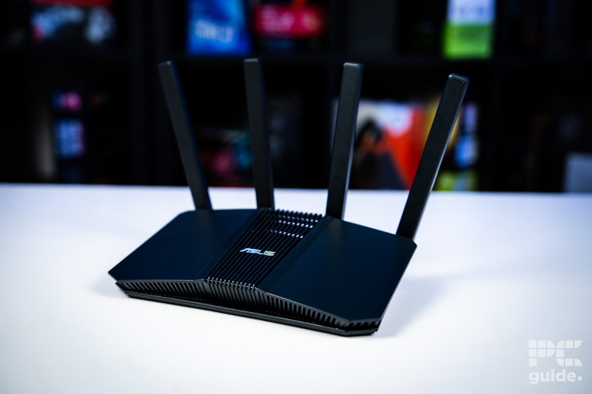 The black ASUS RT-BE58U (BE3600 router sitting on a white table with a background of blurry PC component boxes on a bookshelf.