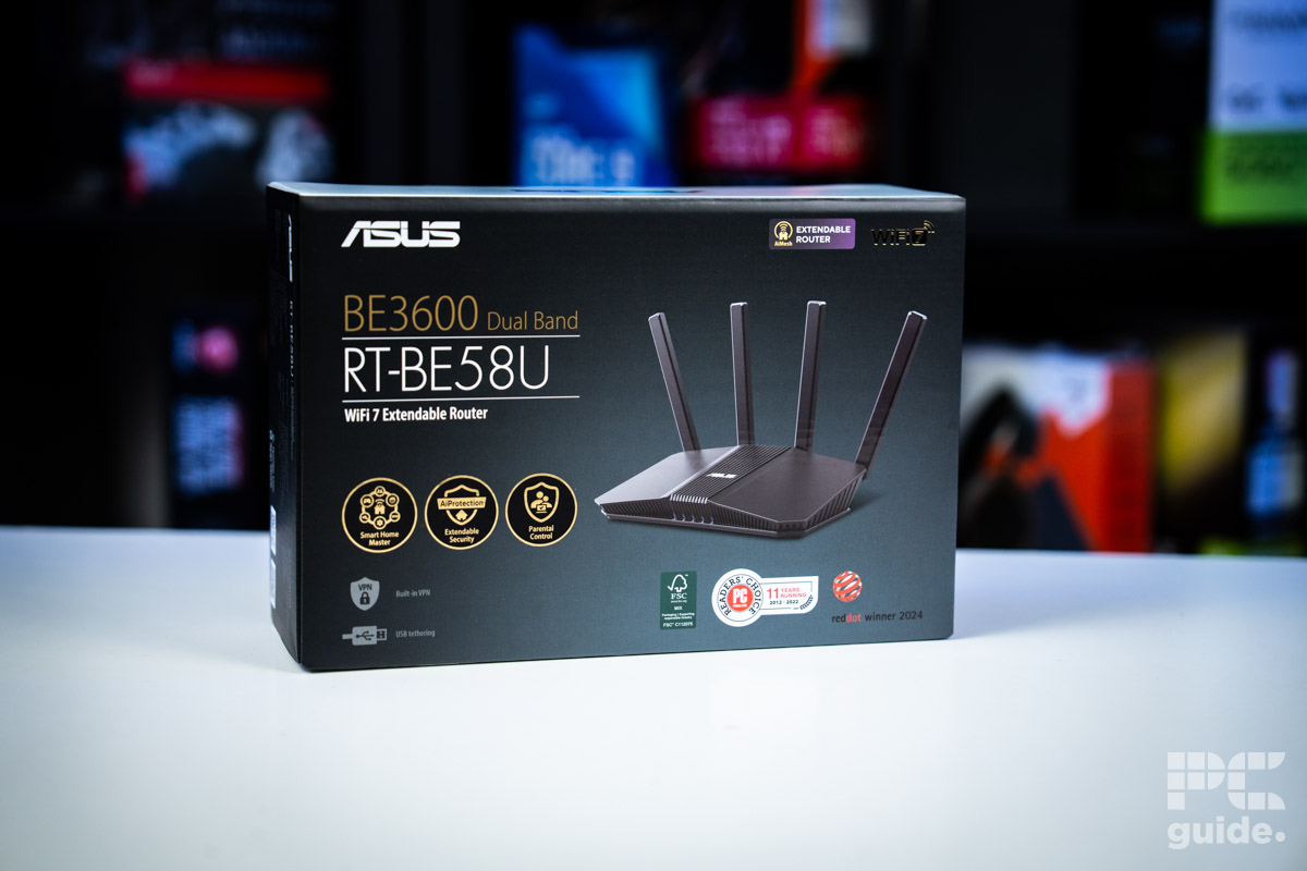 The box for the ASUS RT-BE58U (BE3600 router sitting on a white table with a background of blurry PC component boxes on a bookshelf.